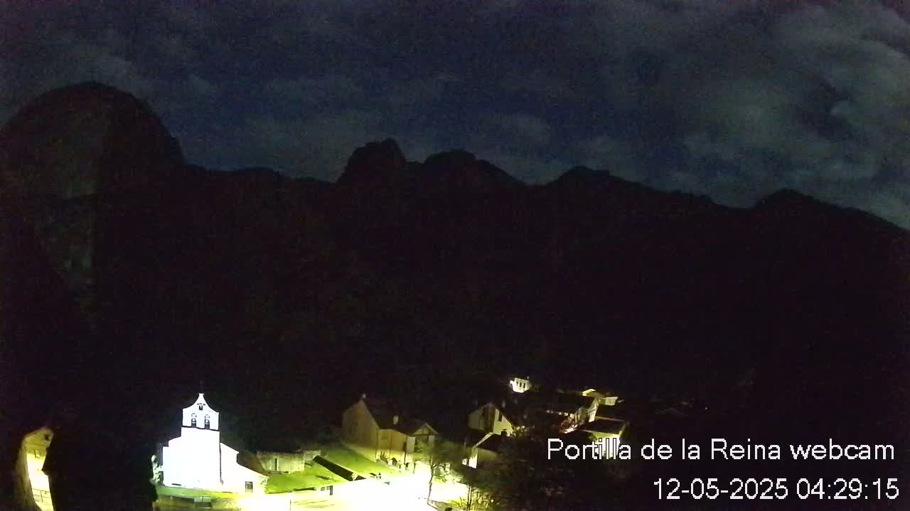 A small village with a brightly lit church is nestled at the base of dark mountains under a partly cloudy night sky.