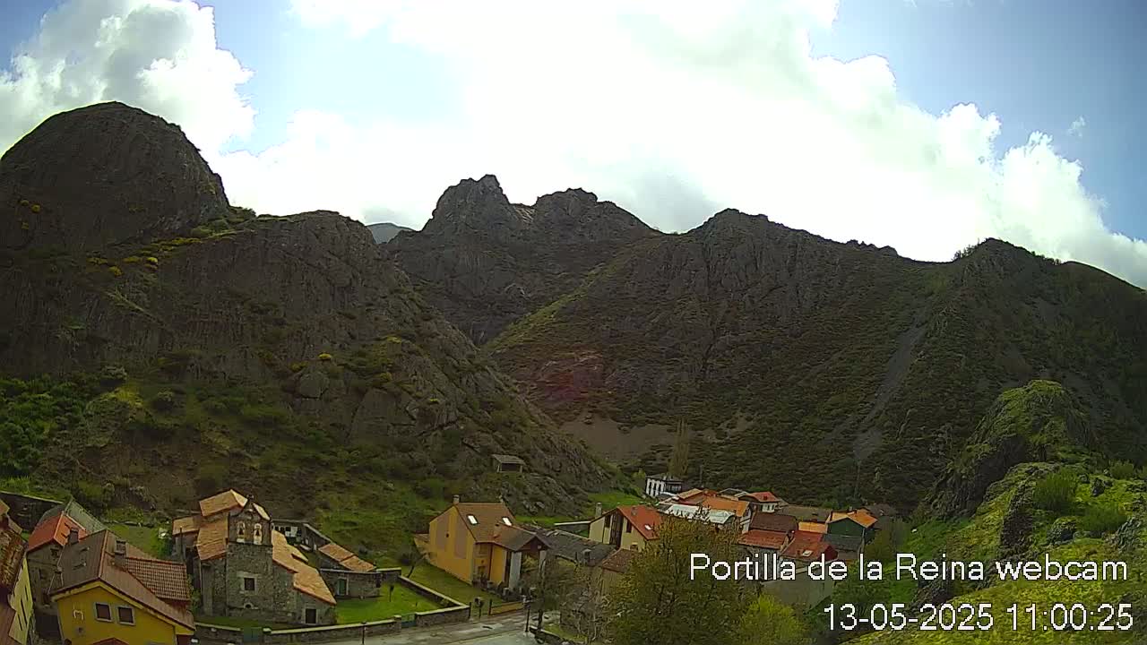 A small village nestled in a valley surrounded by rugged, dark-grey mountains under a partly cloudy sky.