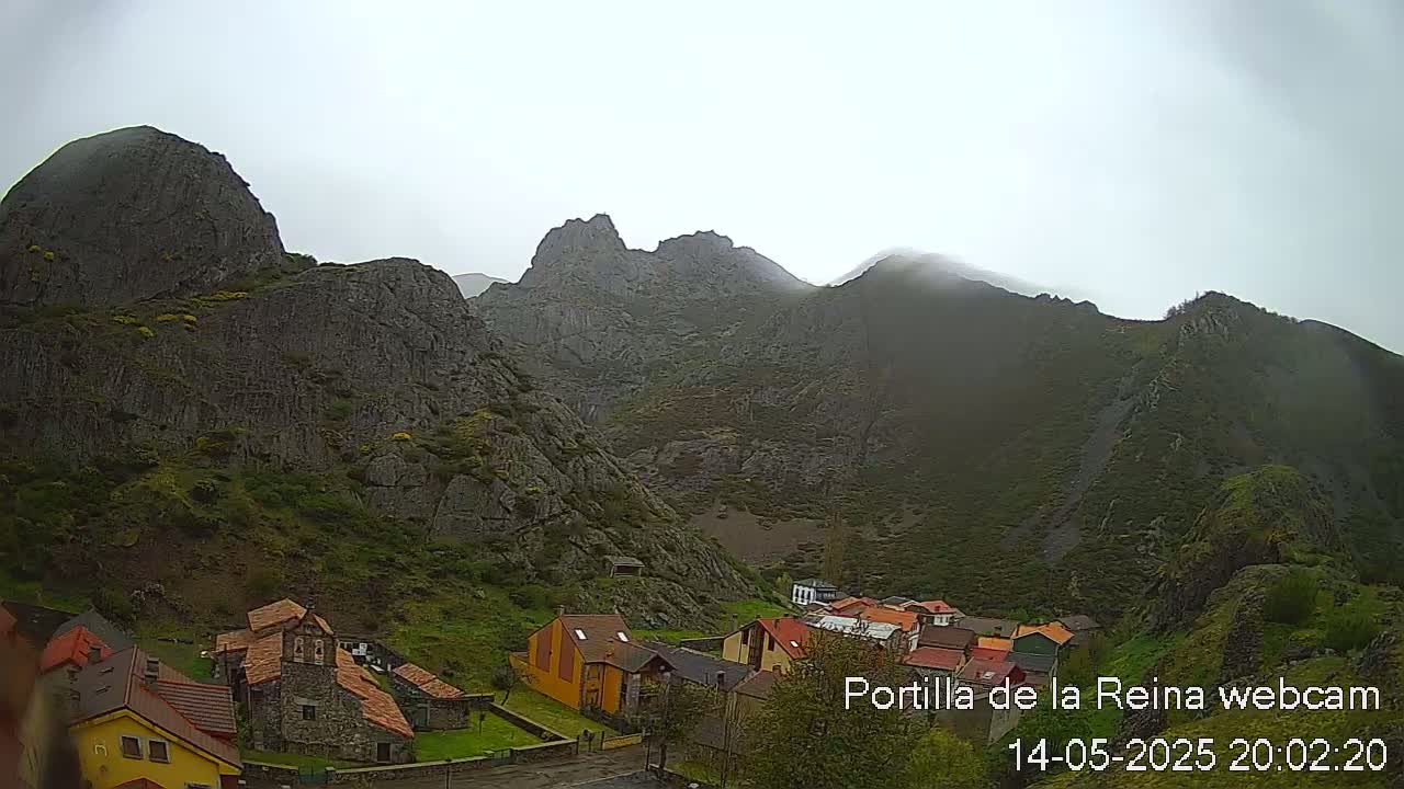 A small village nestled in a valley between imposing, rocky mountains under an overcast sky.