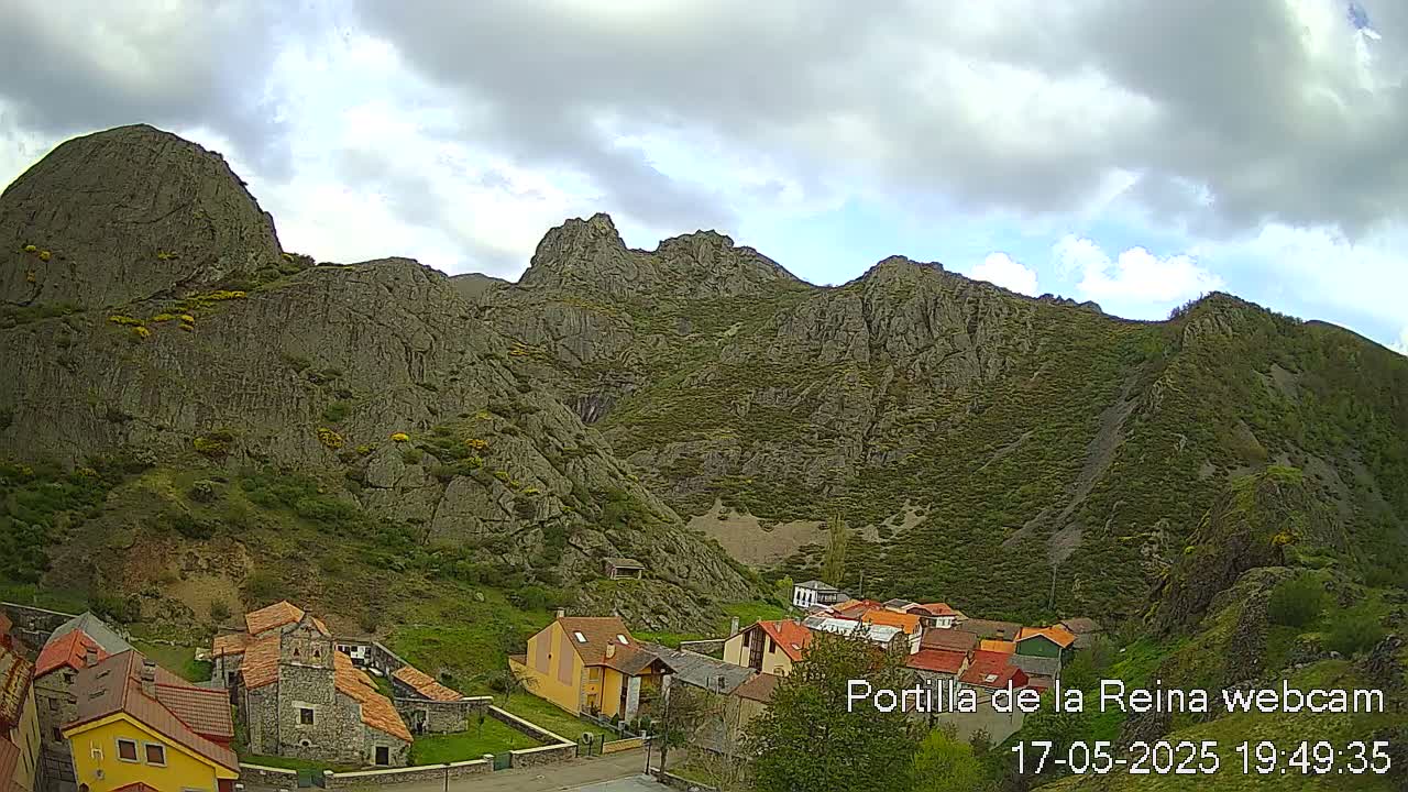 A small village with red-tiled roofs sits at the base of a large, rocky mountain under a partly cloudy sky.
