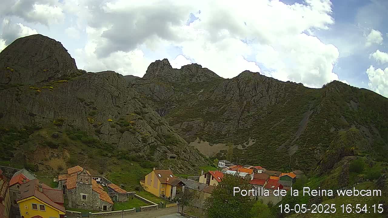 A small village nestled in a valley, surrounded by imposing, rocky mountains under a partly cloudy sky.