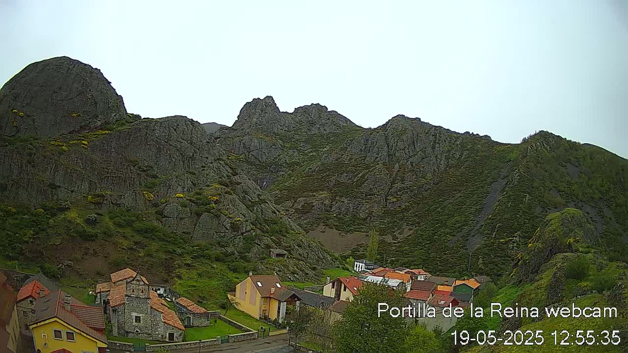 A small village nestled at the base of a large, rocky mountain range under a mostly clear sky.