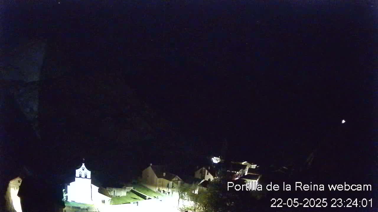 A nighttime, dark view of a small village with a few illuminated buildings under a clear, starlit sky.