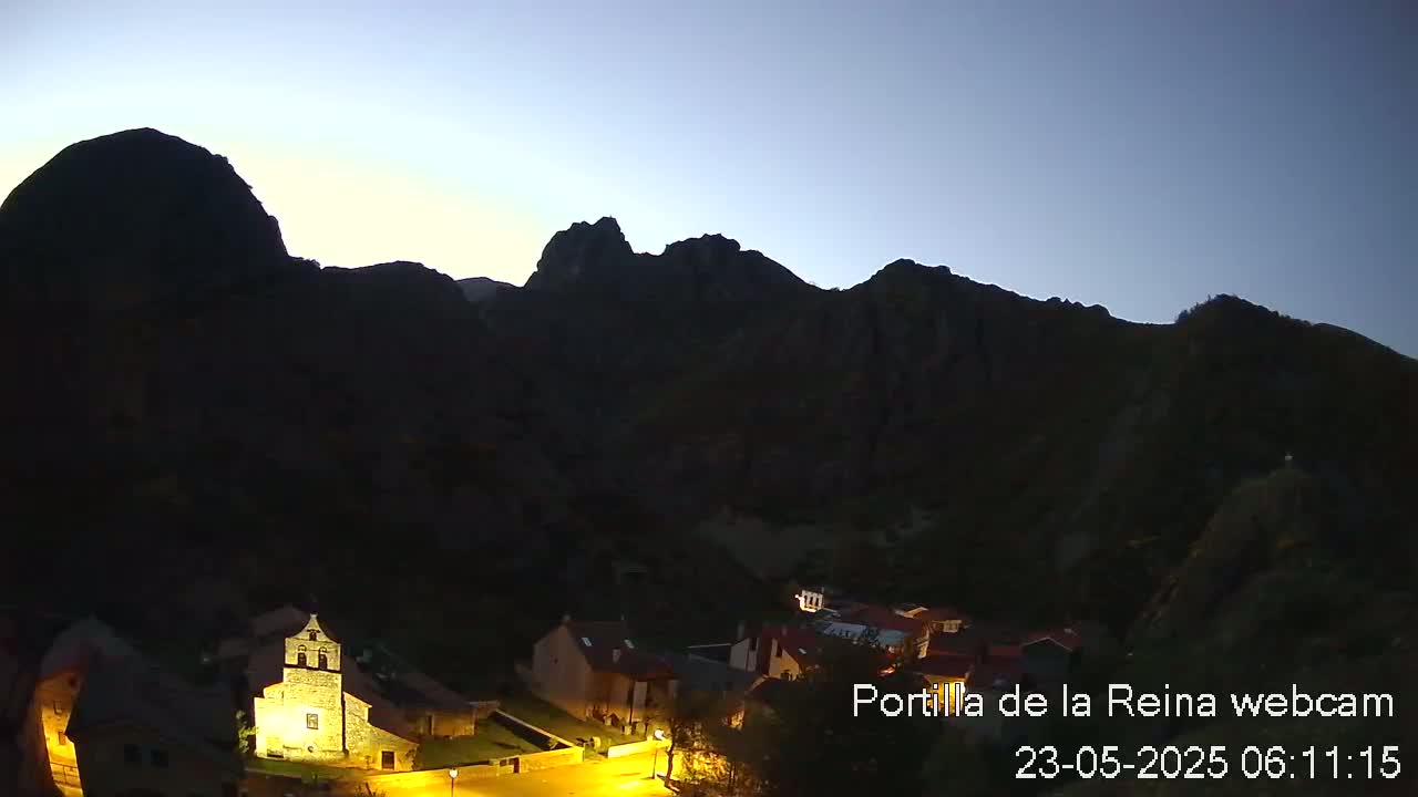 A small village nestled in a valley between dark mountains is illuminated at twilight under a clear, dusky sky.