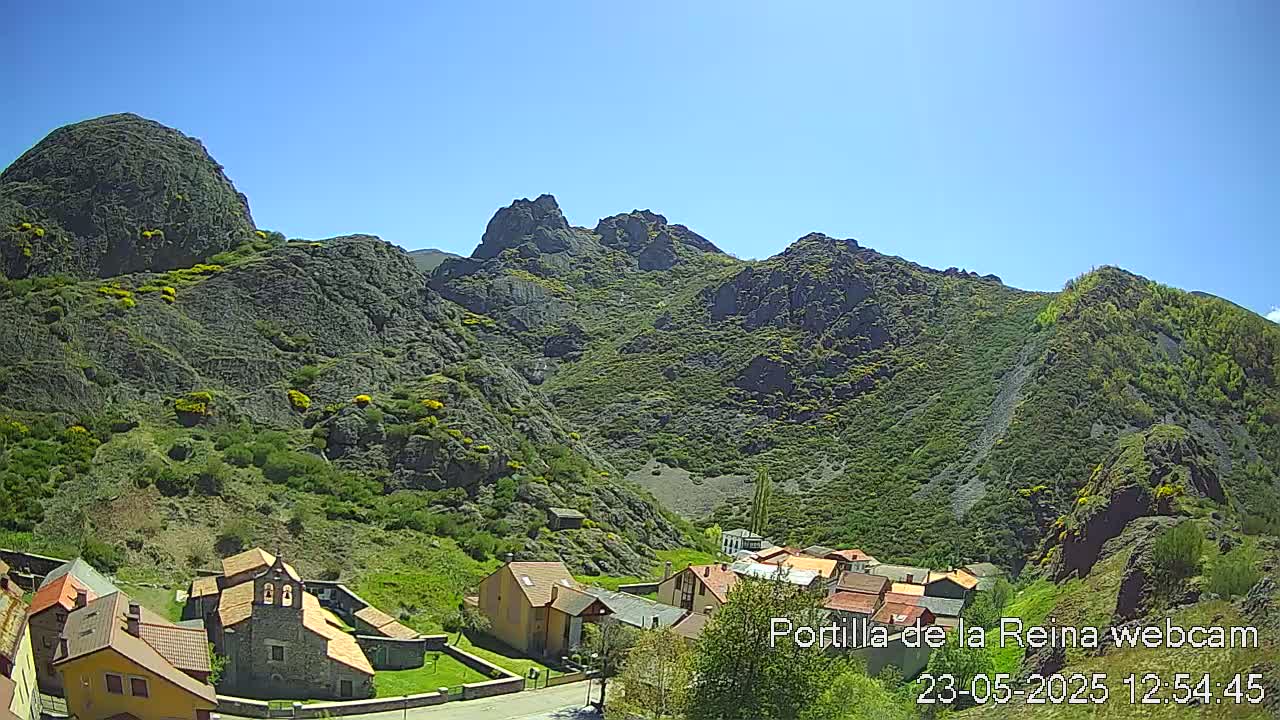 A small village nestled at the base of a rocky, green mountain range under a clear blue sky.