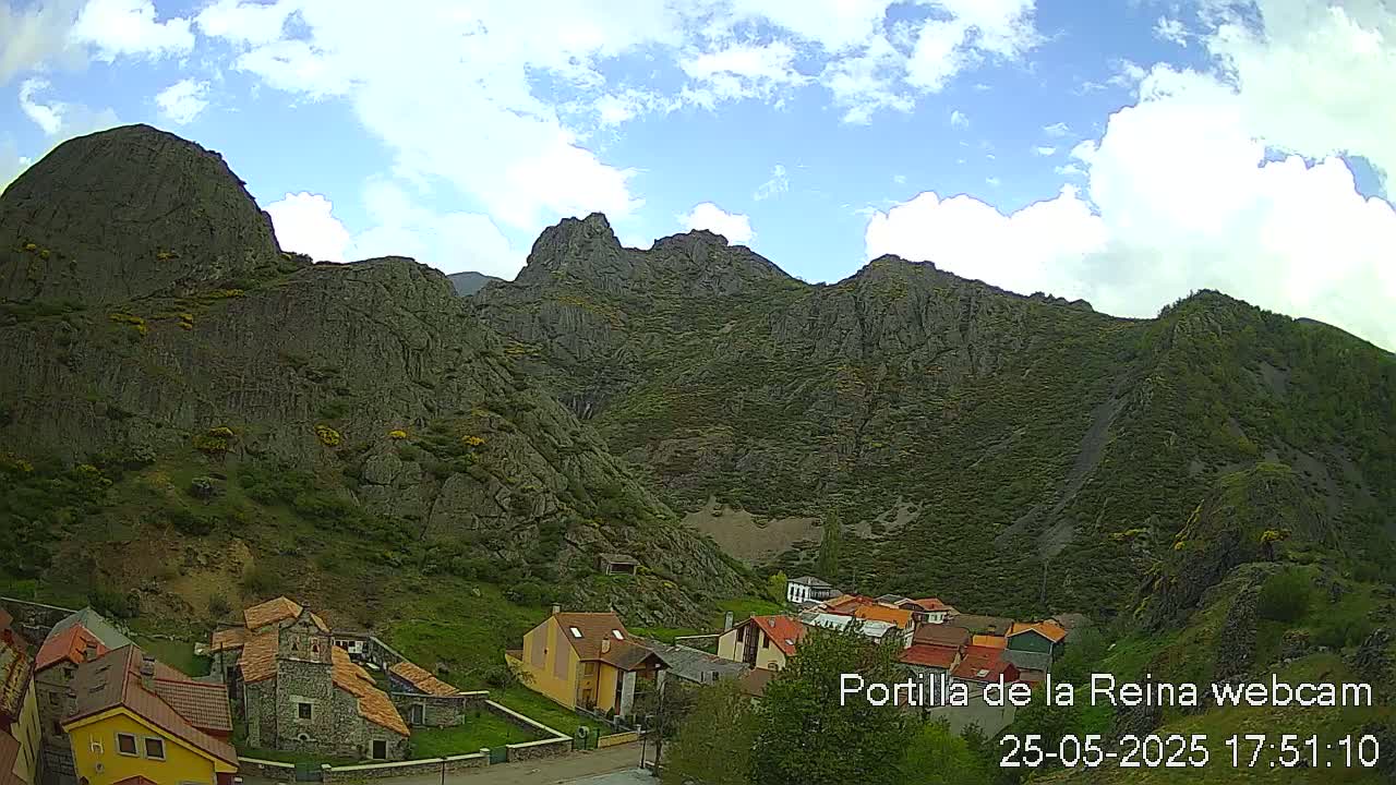 A partly cloudy sky overlooks a small village nestled at the base of a steep, rocky mountain range.