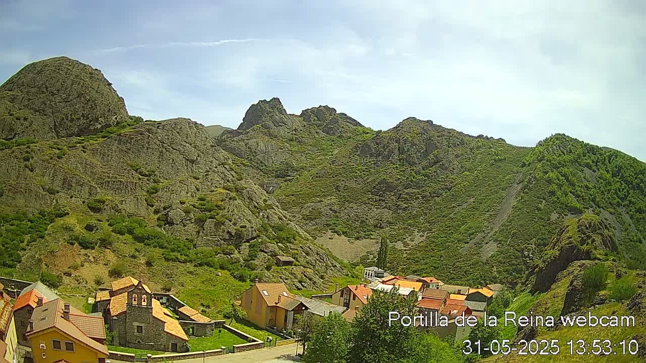 A small village nestled in a valley surrounded by rocky, green-covered mountains under a mostly sunny sky.