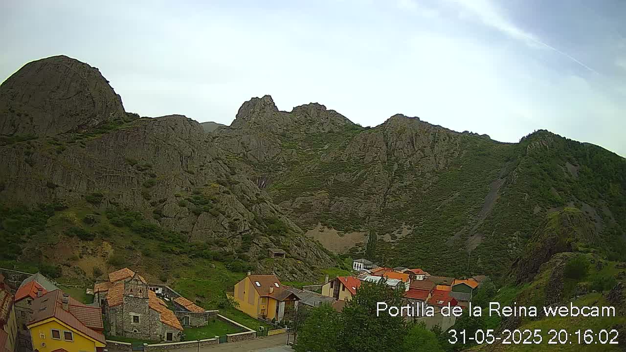 A small village nestled at the base of a large, rocky mountain range under a partly cloudy sky.