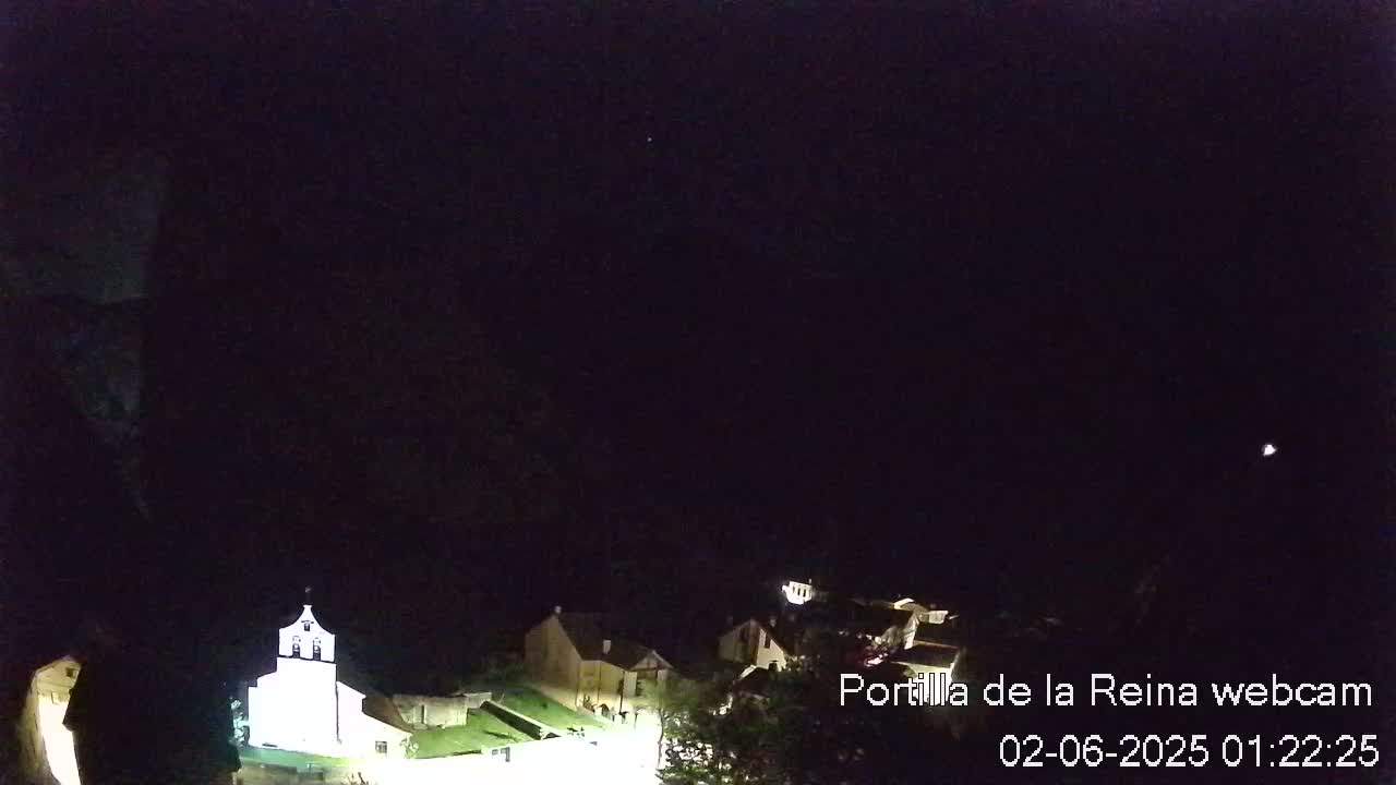 A nighttime view of a small town nestled in a dark landscape, with a church brightly lit in the foreground and a few faint lights visible in the distance under a clear, dark sky.