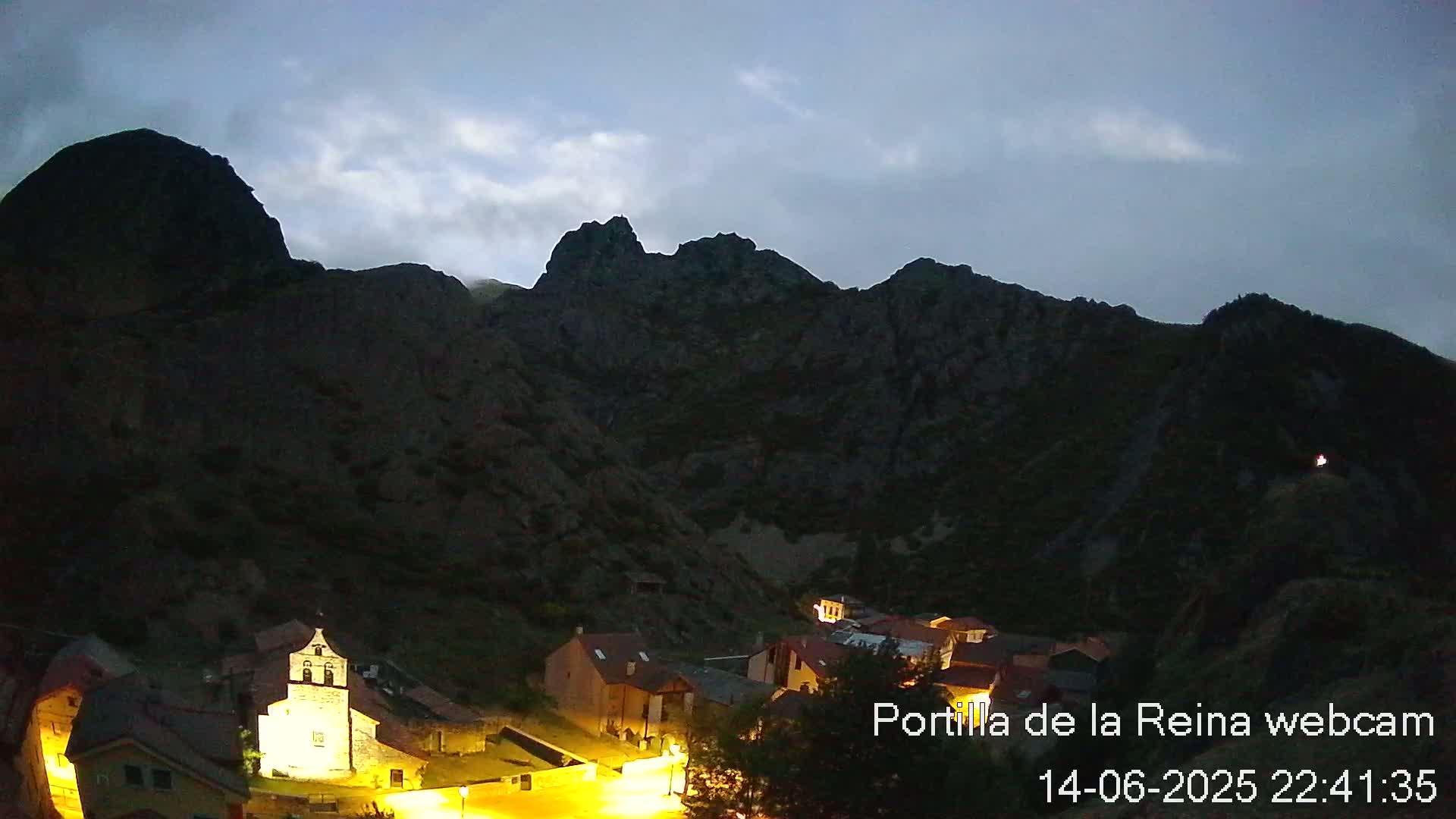 A small village nestled at the base of dark mountains at night, illuminated by a few lights.