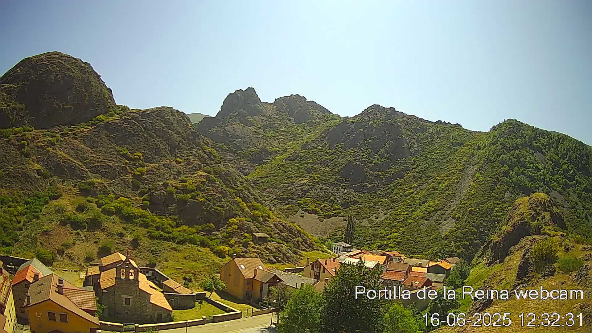 A small village nestled in a valley surrounded by rocky, green-covered mountains under a clear, sunny sky.
