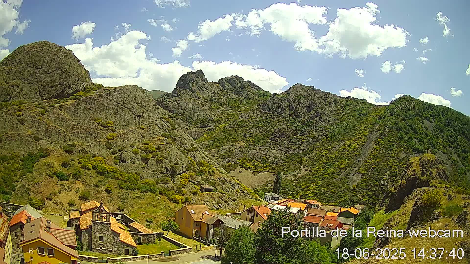 A small village nestled in a valley surrounded by rocky, green-covered mountains under a partly cloudy sky.