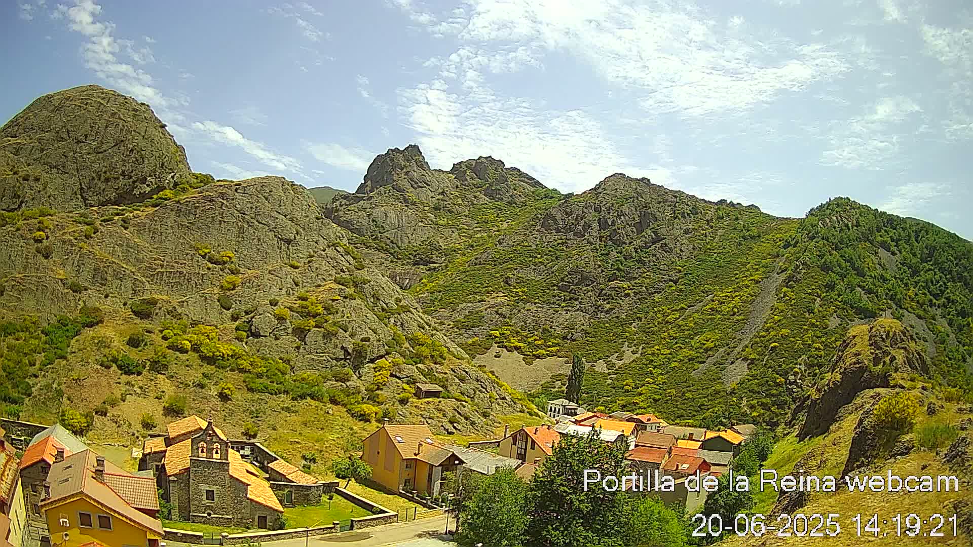 A small village nestled at the base of rocky, green-covered mountains under a mostly sunny sky.