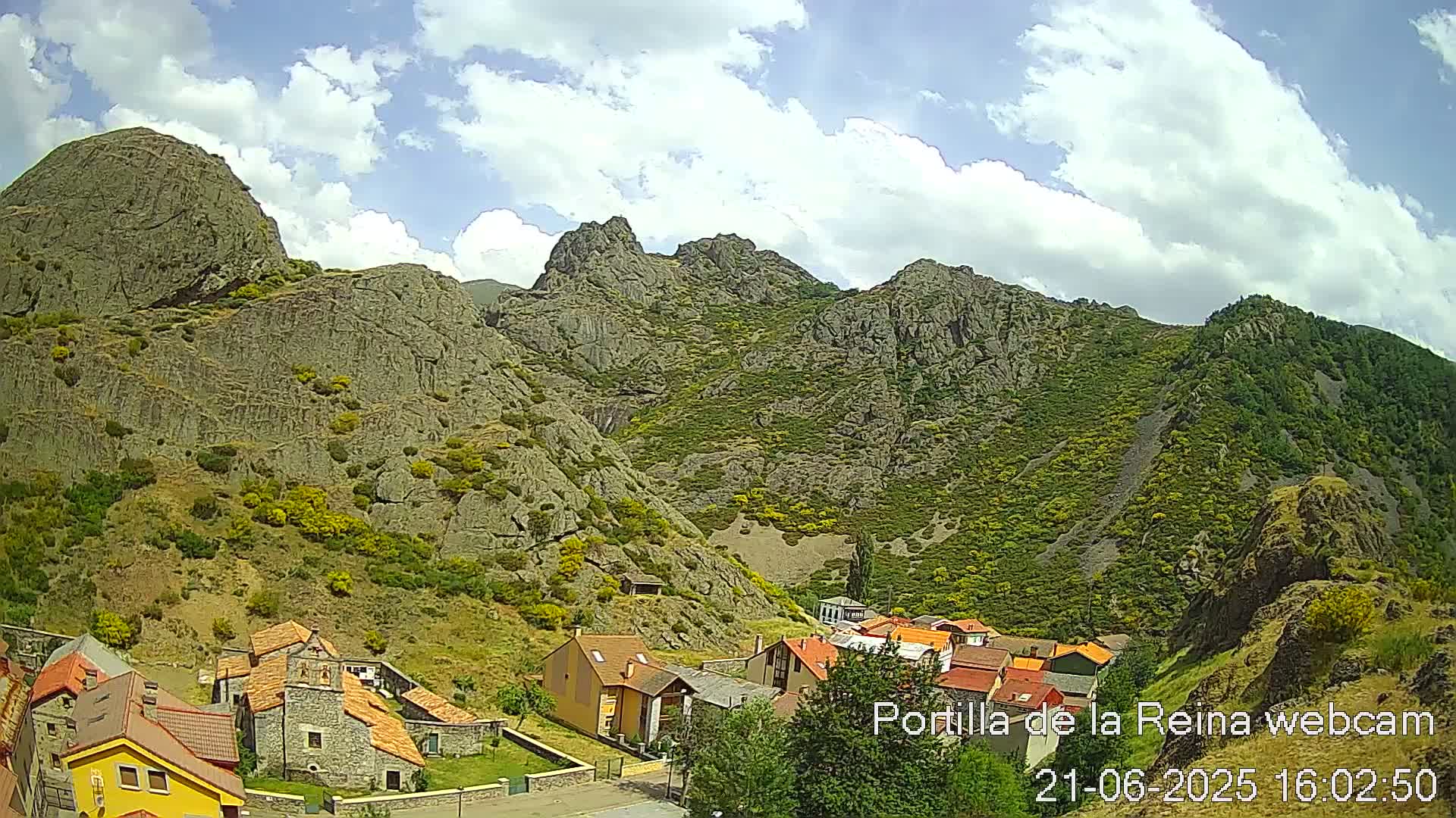 A small village nestled in a valley surrounded by large, rocky mountains under a partly cloudy sky.