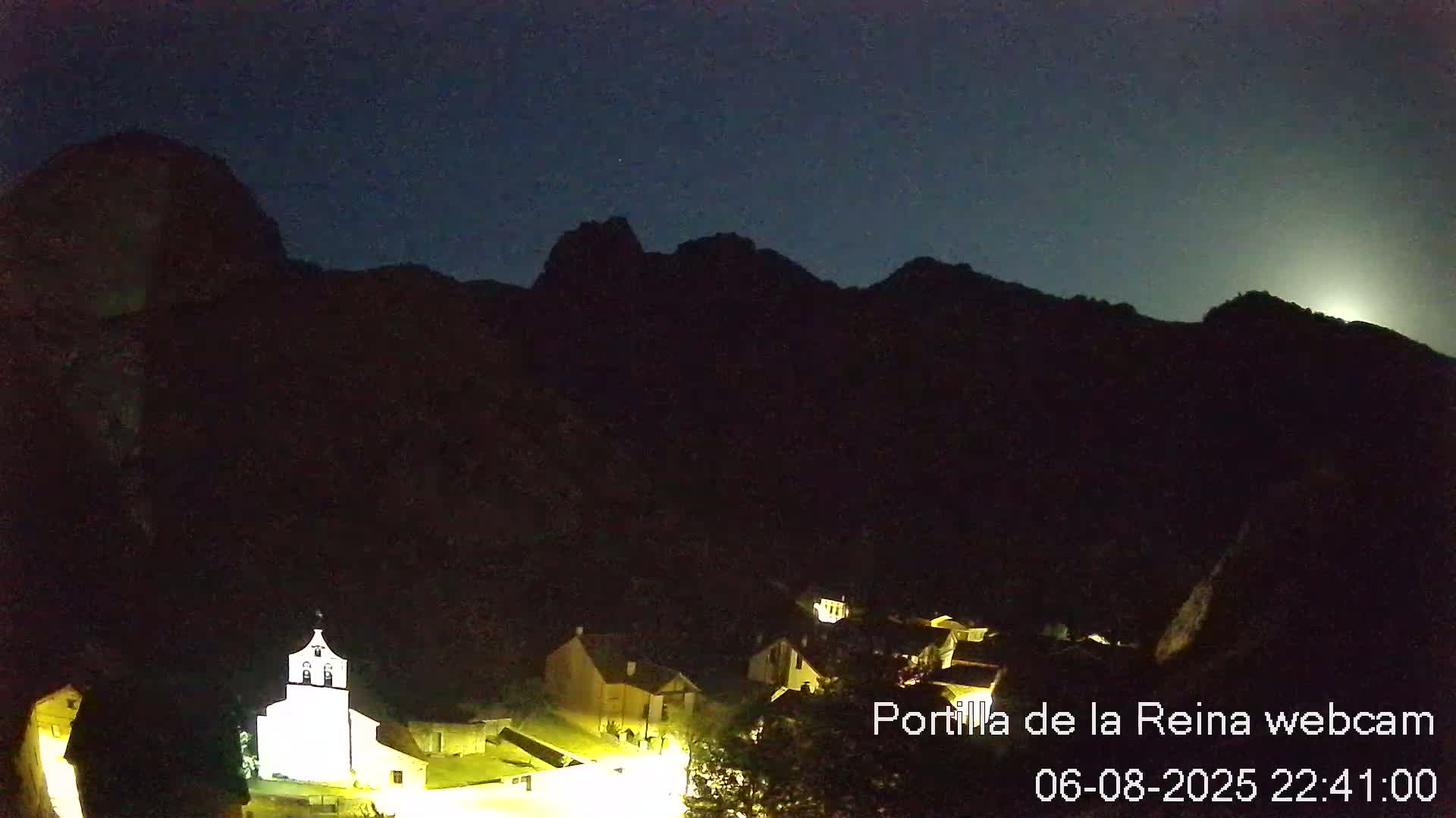 A small, illuminated village nestled at the base of dark mountains under a night sky with a bright moon.
