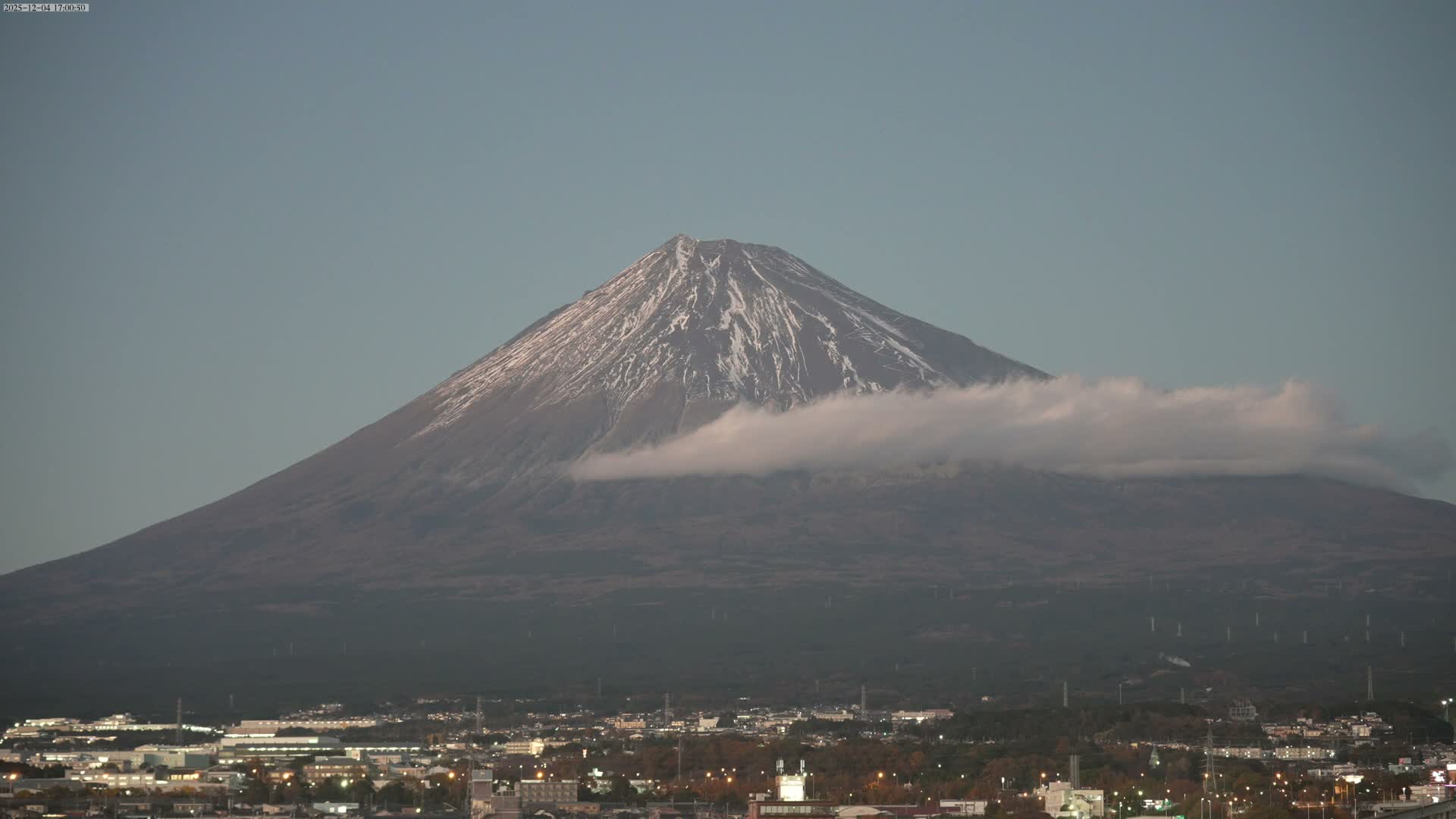 A majestic, snow-capped Mount Fuji, partially encircled by a band of white clouds, looms over a sprawling city with illuminated buildings under a clear, dusk sky.