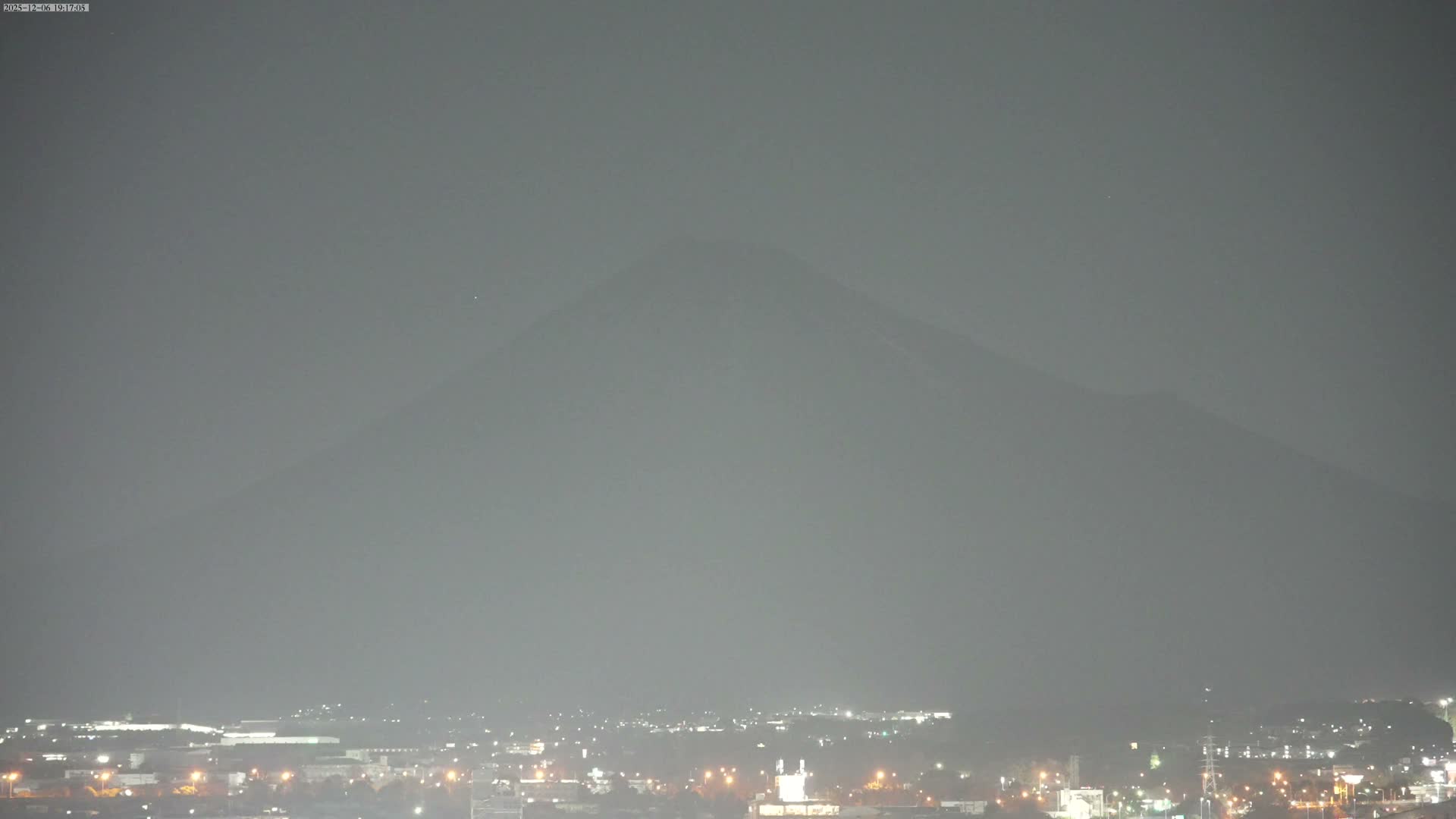 A hazy, dark night reveals the faint silhouette of a large mountain towering over a brightly lit city sprawling across the foreground.
