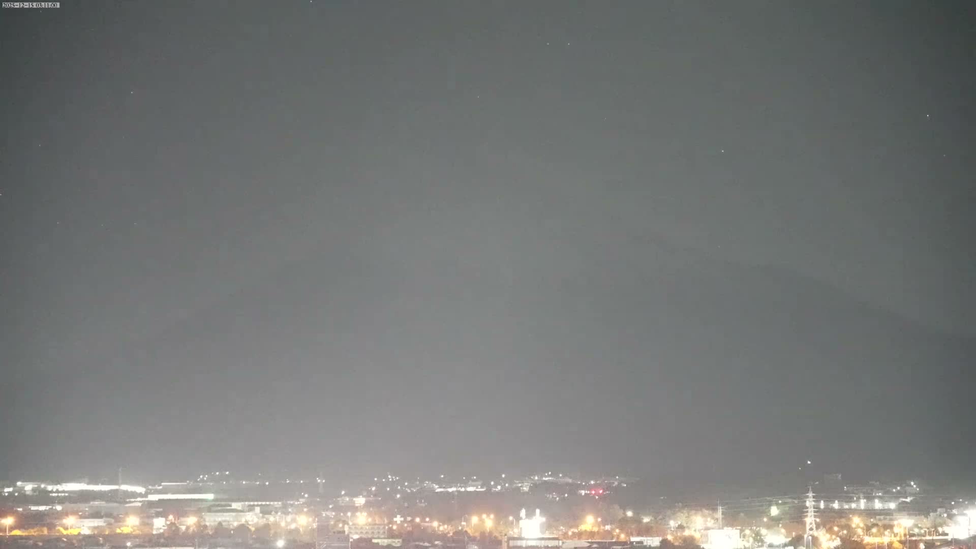 A hazy, dark night reveals the faint silhouette of a large mountain towering over a brightly lit city sprawling across the foreground.