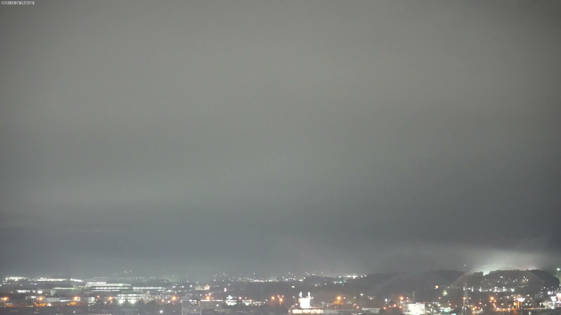 A hazy, dark night reveals the faint silhouette of a large mountain towering over a brightly lit city sprawling across the foreground.