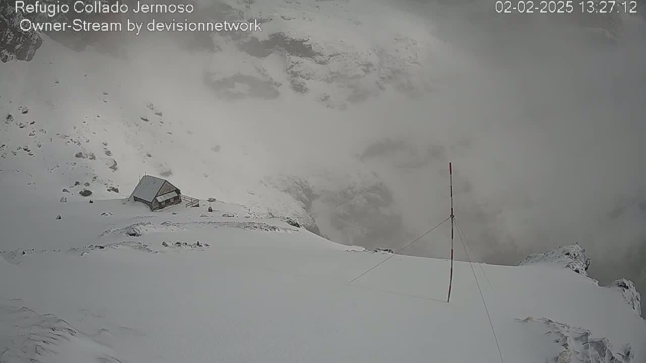 A small, snow-covered cabin sits on a snowy mountainside in foggy conditions.