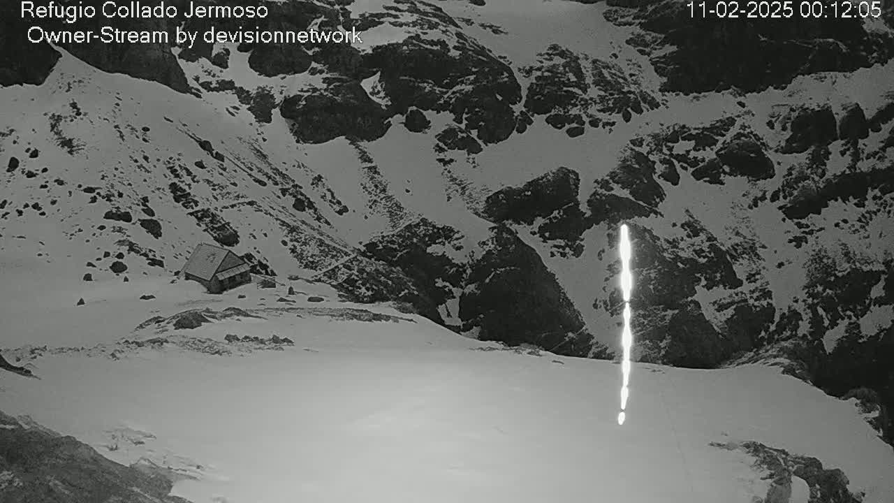 A small structure sits nestled in a snow-covered mountainous area, with a bright vertical streak visible in the snow in the lower right.