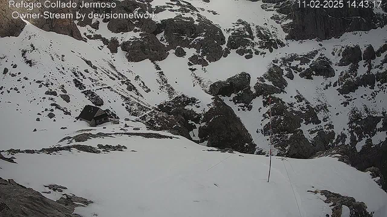 A small building sits nestled in a snowy mountain pass, surrounded by rocky terrain under overcast skies.