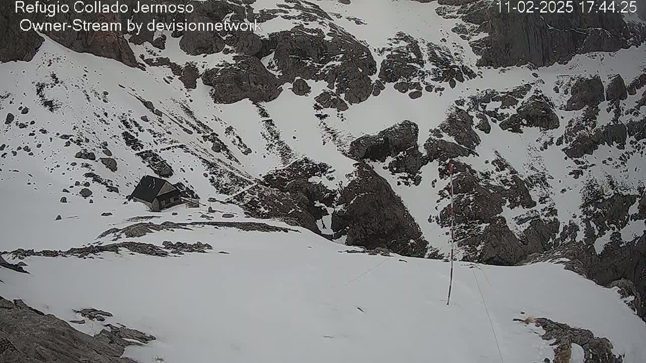 A small cabin sits on a snow-covered mountainside, overlooking a rocky valley also covered in snow.