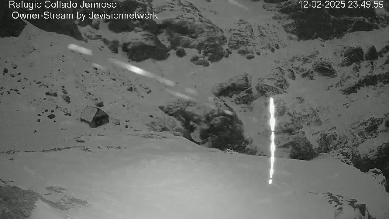 A nighttime, snowy, mountainous landscape shows a small structure nestled in the snow, with streaks of light possibly from snow or ice falling in the distance.