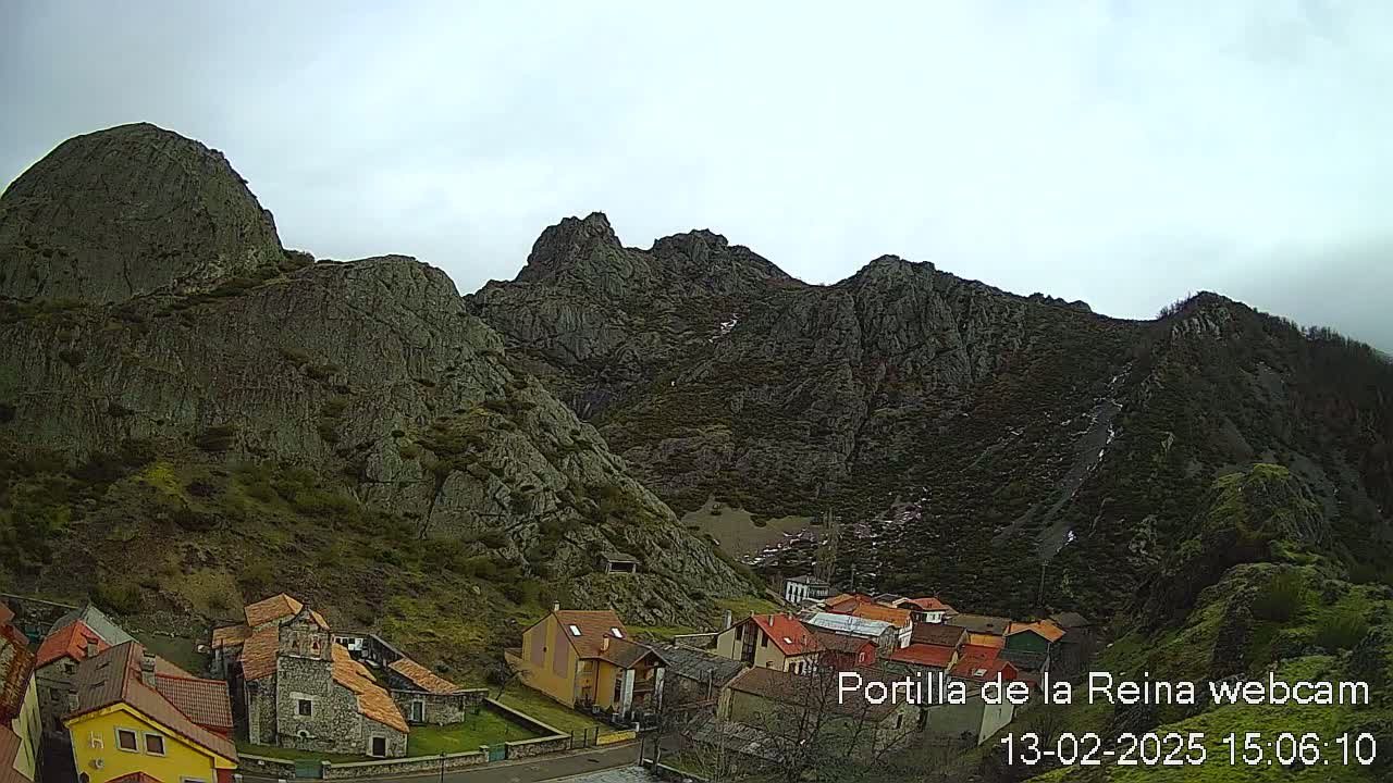 A small village nestled at the base of large, dark, rocky mountains under an overcast sky.