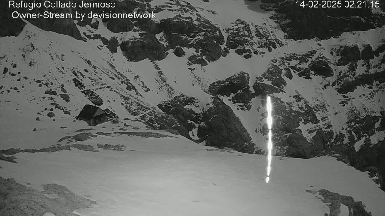 A nighttime, snow-covered mountainous landscape shows a small building nestled into the side of a hill, with a bright vertical streak of light in the foreground.