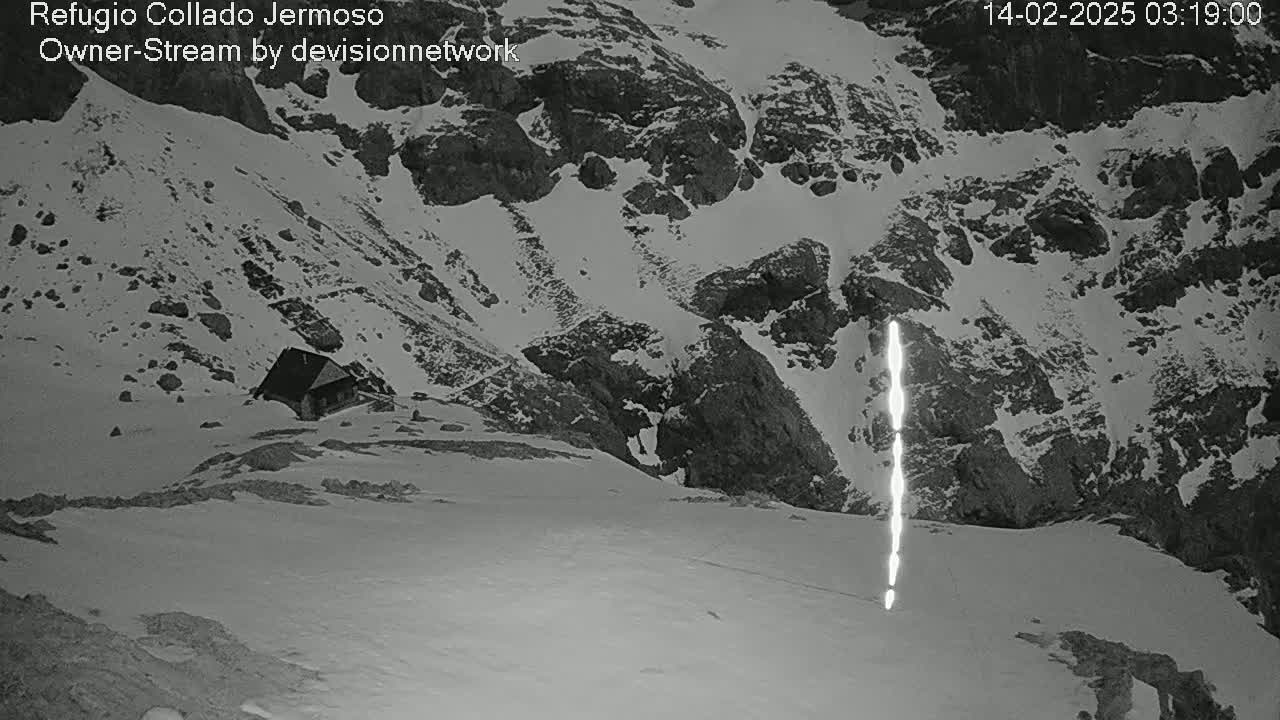 A small cabin sits nestled in a snow-covered mountainous area at night, with a bright vertical streak visible in the snow.