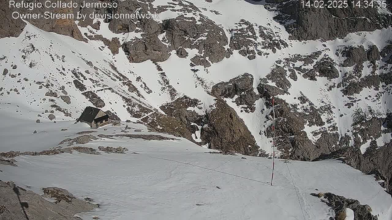 A small cabin sits nestled in a snow-covered mountain pass on a sunny day.