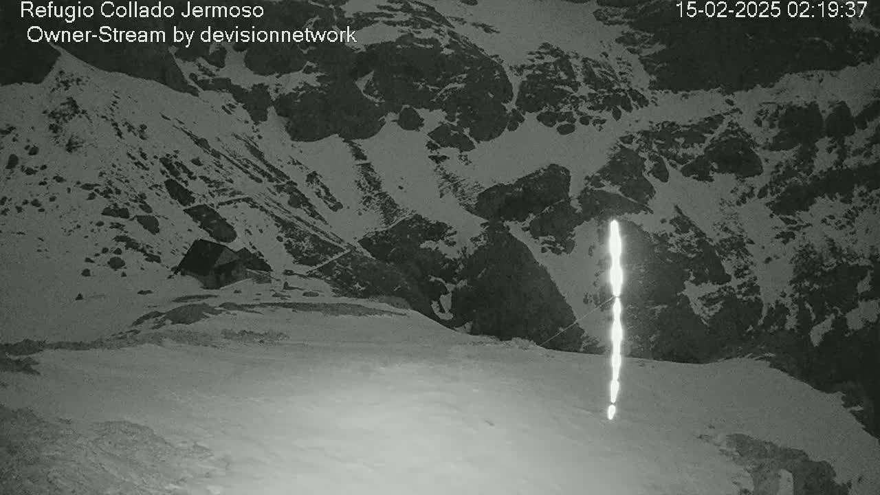 A nighttime, snow-covered mountain landscape with a small building visible in the distance and a vertical bright light source in the foreground.