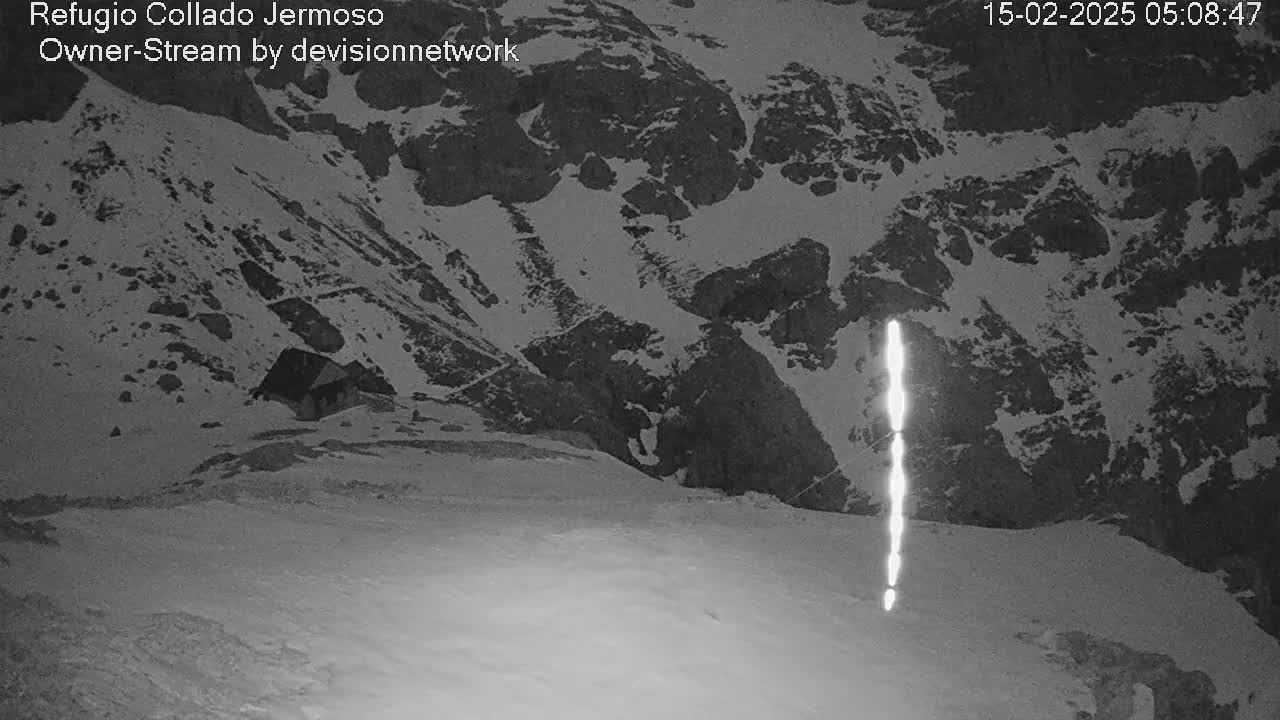 A nighttime, snow-covered mountain landscape shows a small building nestled in a valley, with a bright vertical streak of light in the foreground.
