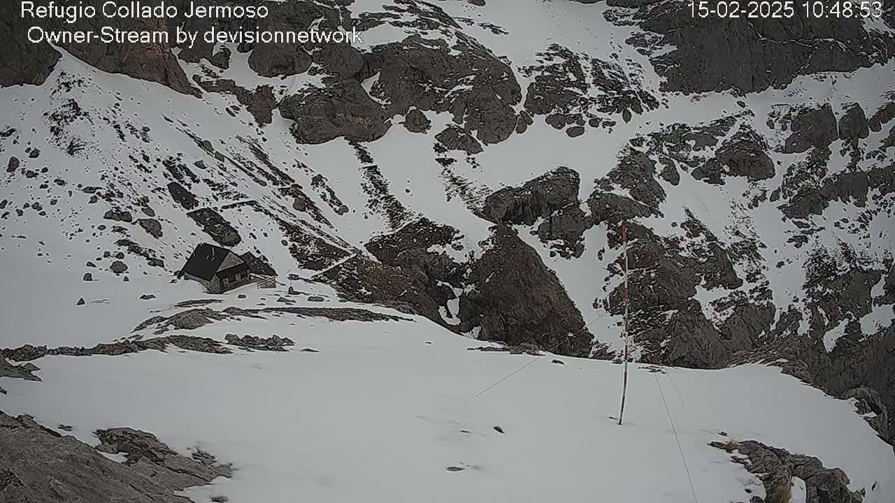 A small building sits nestled in a snowy mountain valley, with rocky outcrops and a thin line stretching across the snow-covered terrain.