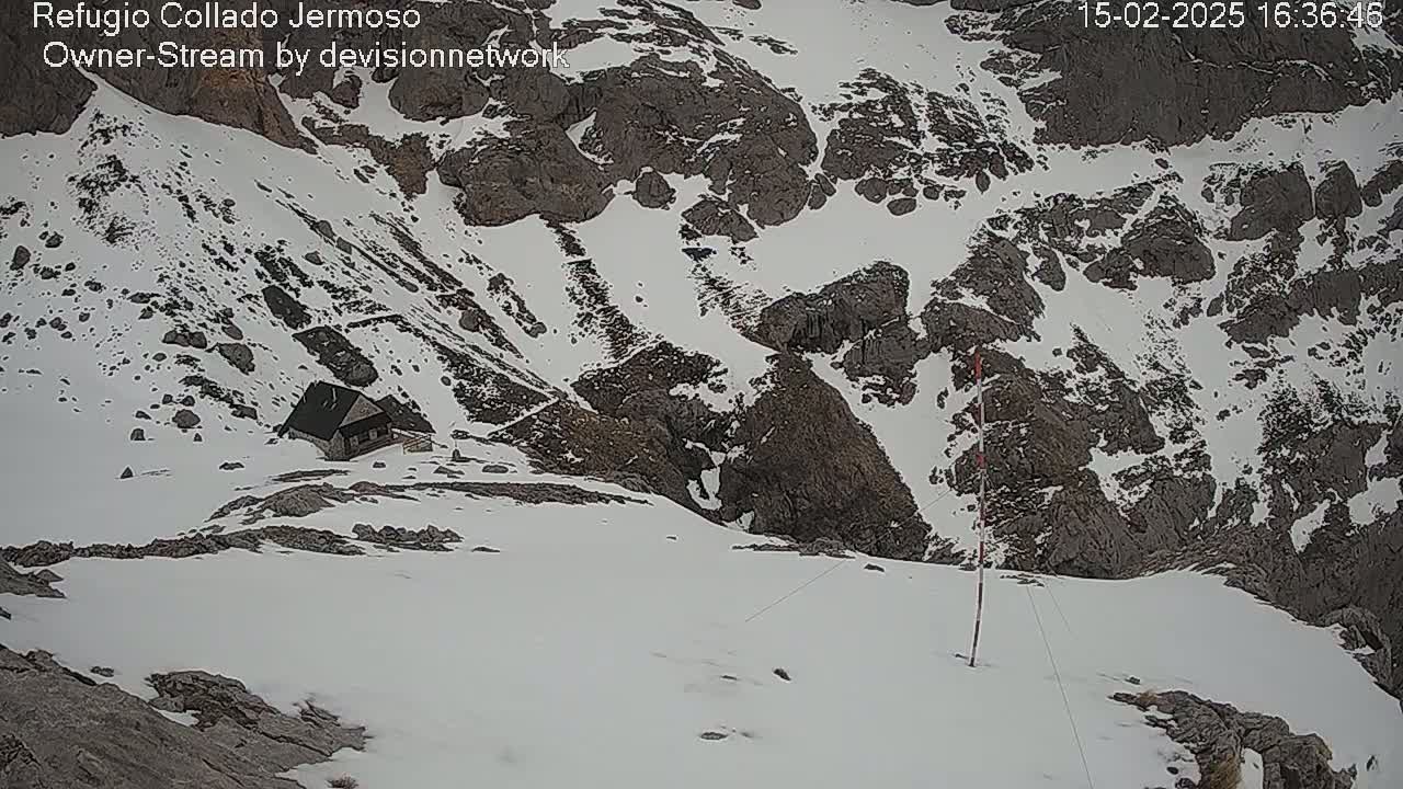 A small cabin sits nestled in a snowy mountain pass, with rocky outcrops visible under a layer of snow.