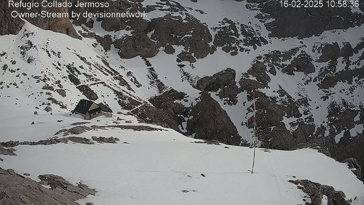 A small cabin sits on a snow-covered mountainside, overlooking a rocky valley also covered in snow.