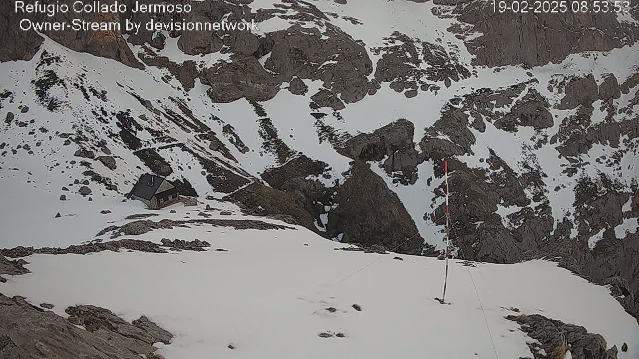 A small cabin sits nestled in a snow-covered mountain pass, with rocky outcrops and patches of snow visible on the surrounding landscape.