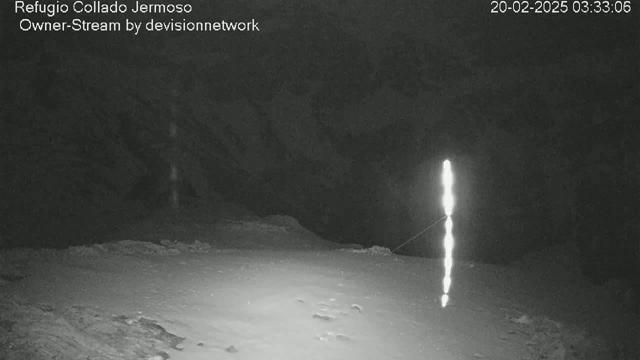A nighttime, snow-covered landscape is illuminated by a vertical line of bright lights extending from the ground to near the top of a dark, rocky outcropping.