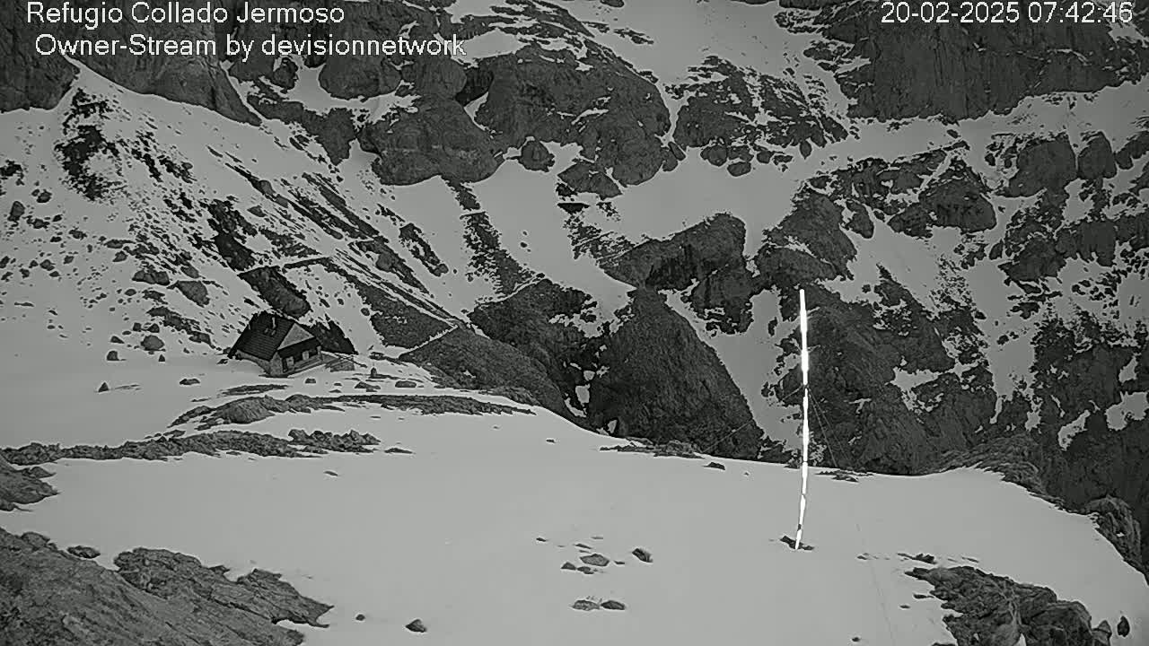 A small cabin sits nestled in a snow-covered mountainous region, with a tall, thin, light-colored pole standing in the foreground.