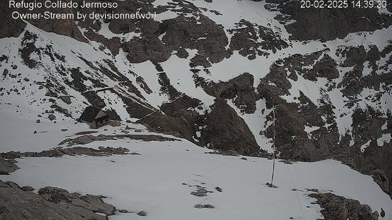 A small cabin sits nestled in a snowy, rocky mountain pass.