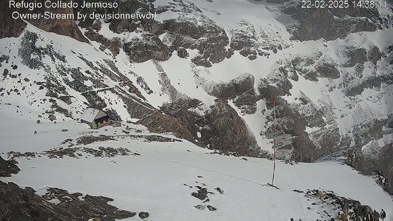 A small cabin sits on a snow-covered mountainside, overlooking a rocky, snow-filled valley in snowy conditions.