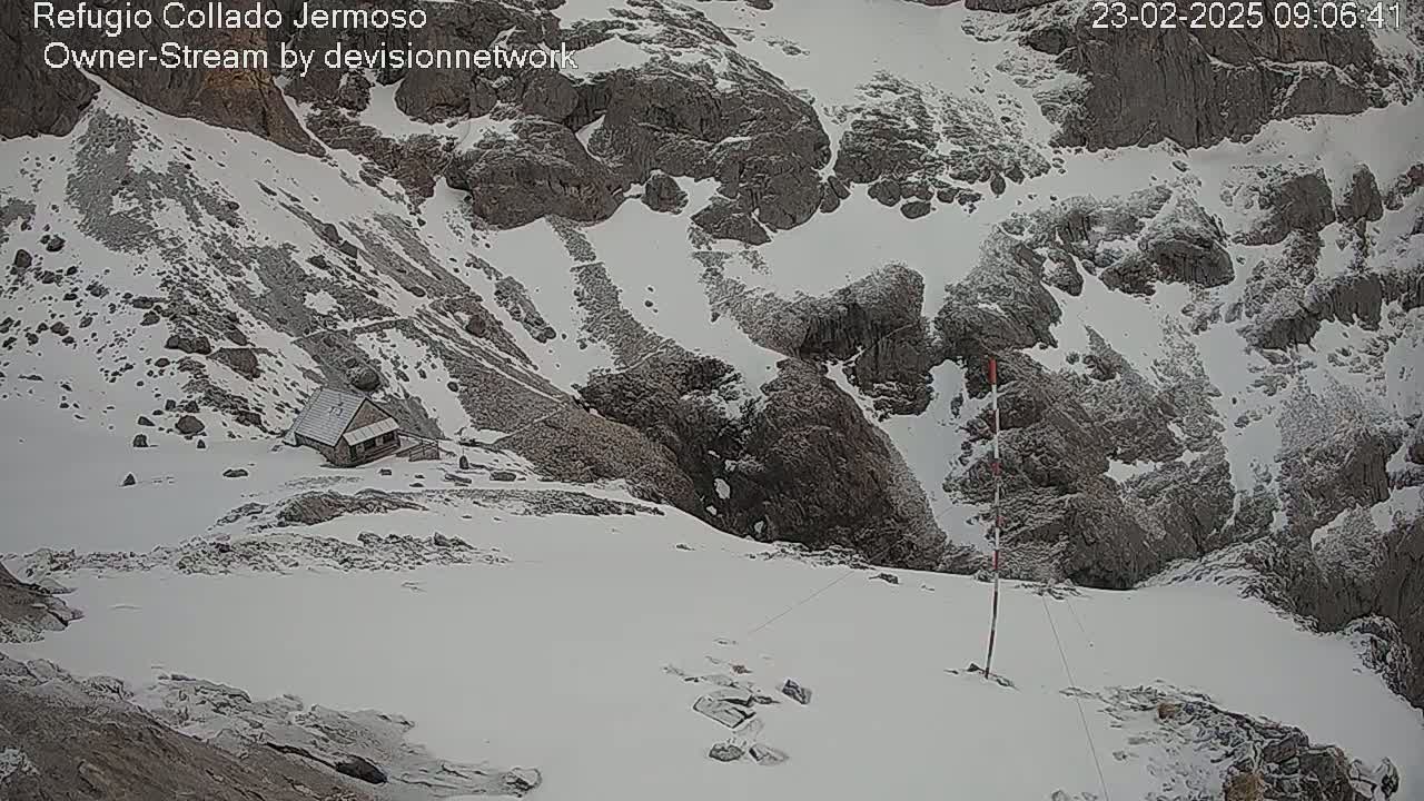 A small cabin sits on a snow-covered mountainside amidst rocky terrain under an overcast sky.