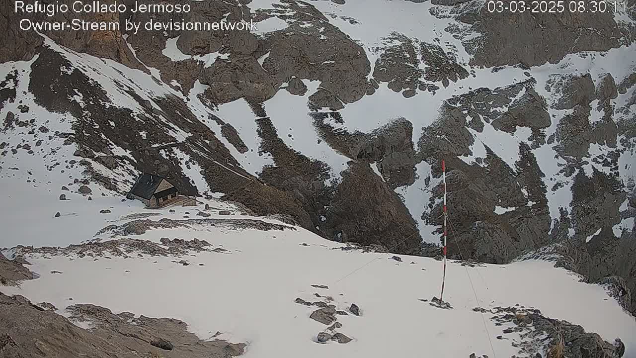 A small cabin sits nestled in a snow-covered mountainous region, with rocky outcrops and patches of snow visible in the background under a cloudy sky.