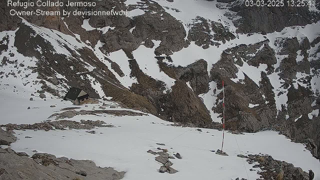 A small cabin sits nestled in a snowy, rocky mountain pass.
