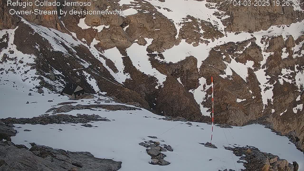 A snow-covered mountain landscape with a small building nestled into a rocky slope, under partly sunny conditions.