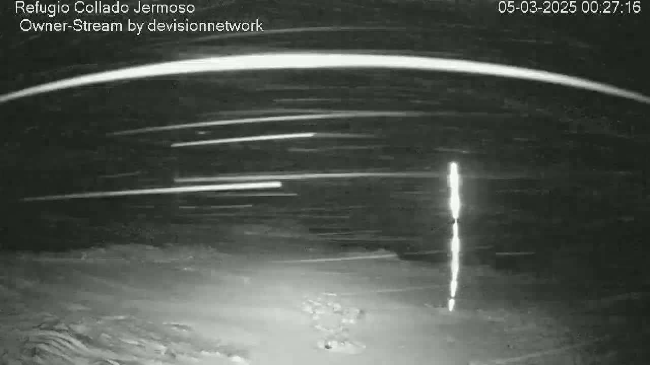 A nighttime, snowy scene shows several streaks of light in the sky, possibly from car headlights or meteors, along with a tall, bright vertical light source in the lower-right.