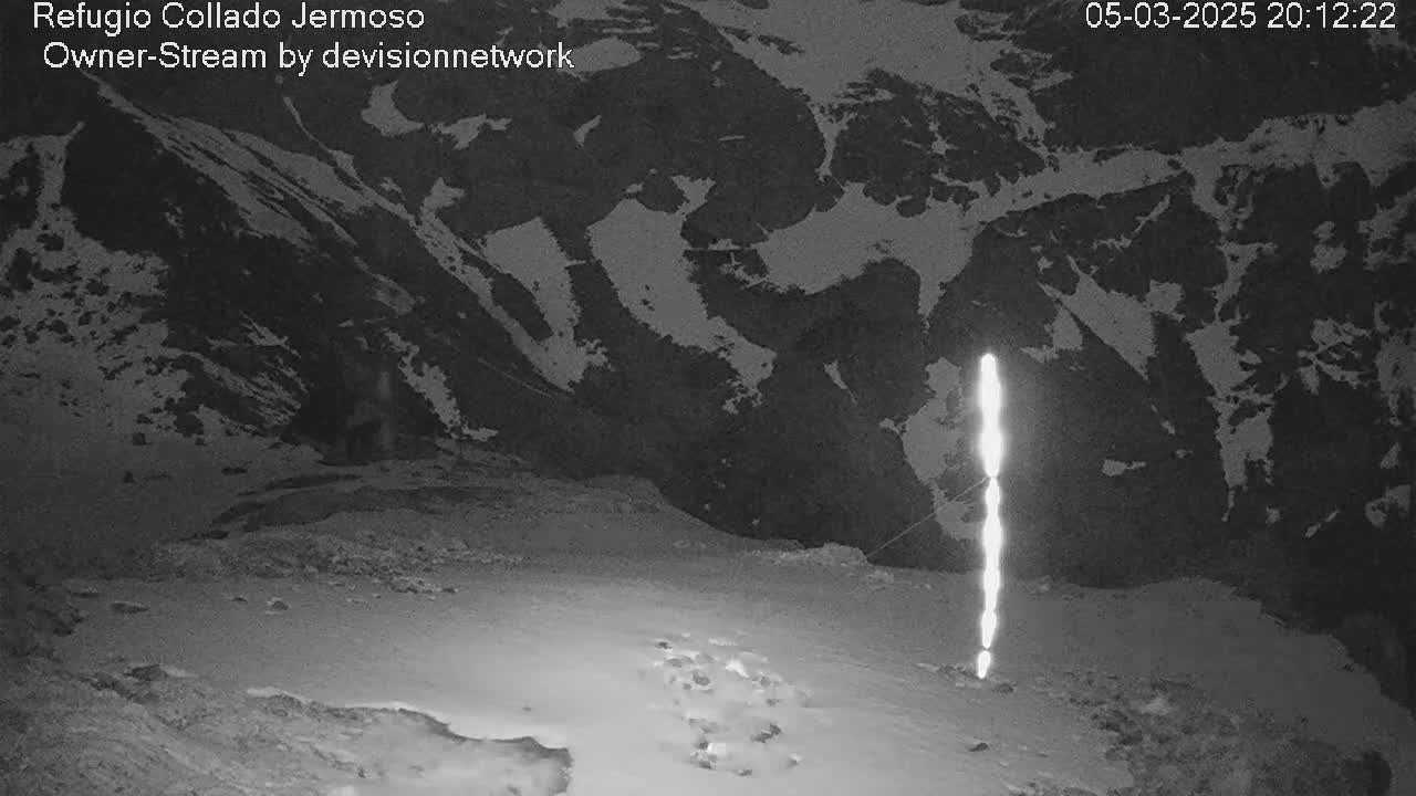 A bright vertical light source stands in a snow-covered mountainous area at night.