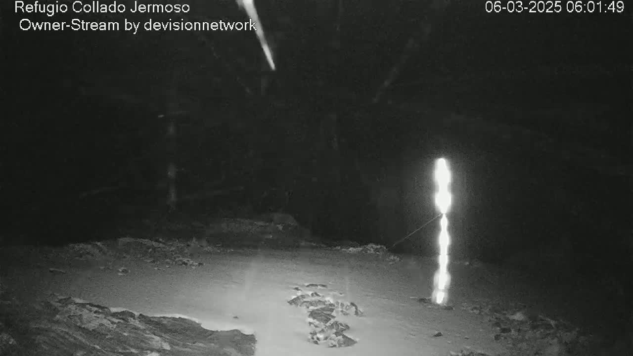 A nighttime, snowy scene shows a tall, vertical light source in the center, with a faint streak of light in the upper left corner.