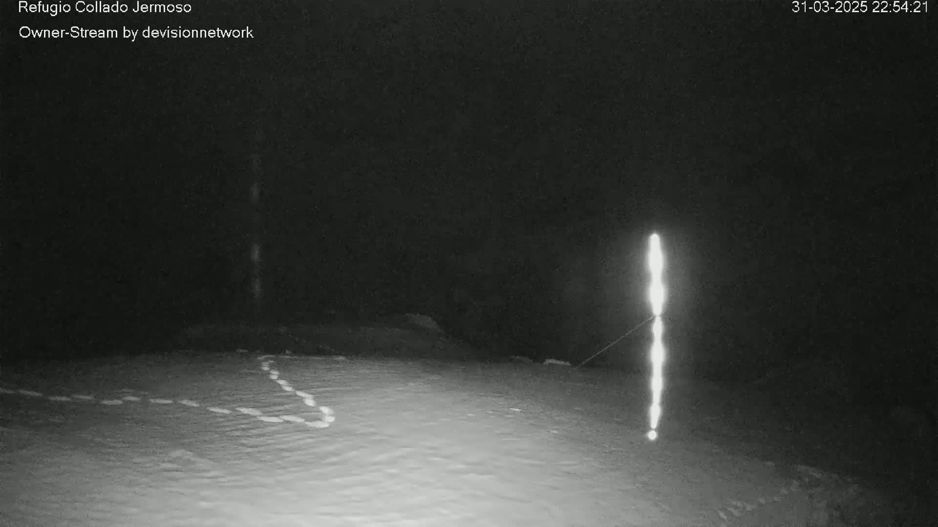 A nighttime, snow-covered scene shows a bright vertical light source and faint animal tracks in the snow.